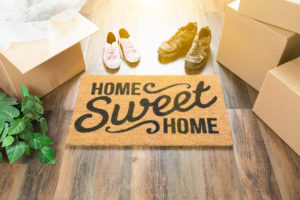 Home Sweet Home Welcome Mat, Moving Boxes, Women and Male Shoes and Plant on Hard Wood Floors. Home Sweet Home Welcome Mat, Moving Boxes, Women and Male Shoes and Plant on Hard Wood Floors.
